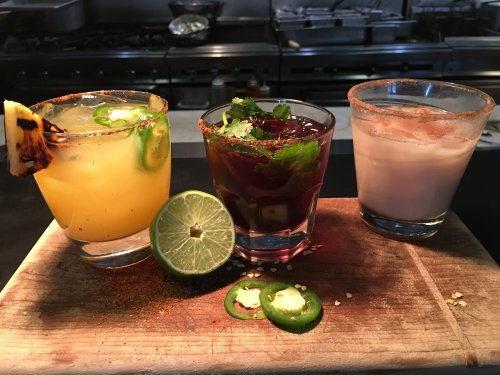 Three colorful cocktails on a wooden board: a yellow drink with jalapeños, a dark drink with cilantro, and a creamy beverage.