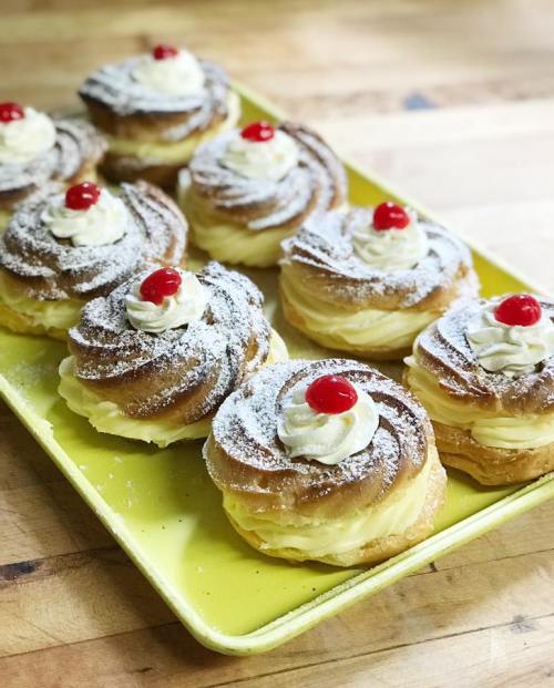 A platter of cream-filled pastries topped with whipped cream and cherries, dusted with powdered sugar.