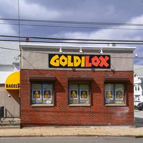 A brick building with a sign reading "GOLDILOX" and a yellow awning labeled "BAGELS." Windows are covered with posters.
