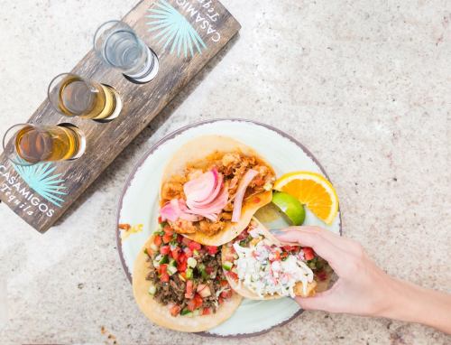 A hand reaches for a taco on a plate with two more tacos, garnished with fresh toppings, alongside a flight of tequila.