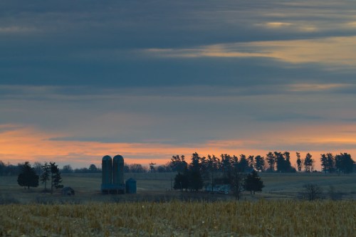 A serene rural landscape at sunset, featuring silos and trees against a colorful sky.