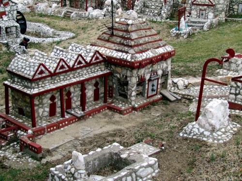 A miniature village featuring stone buildings with red accents, surrounded by grassy areas and small structures.