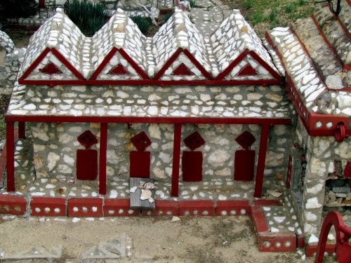 A miniature stone building with a red-trimmed roof and decorative windows, set in a landscaped area.