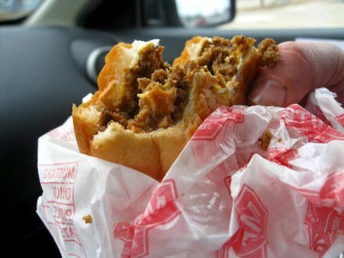 A hand holding a partially eaten sandwich wrapped in red and white paper, inside a car.