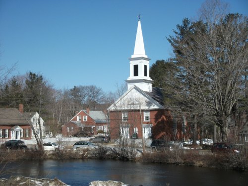 A white steeple church beside a river, with snow on the ground and houses in the background under a clear blue sky.