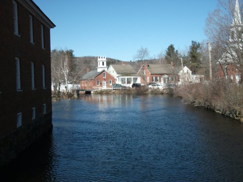 A serene river scene with brick buildings and a church in the background, surrounded by trees under a clear blue sky.