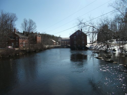 A serene river scene with historic buildings on the banks, surrounded by trees and a clear blue sky.