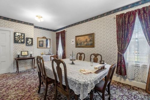 A vintage dining room with floral wallpaper, a lace tablecloth, wooden chairs, and decorative curtains.