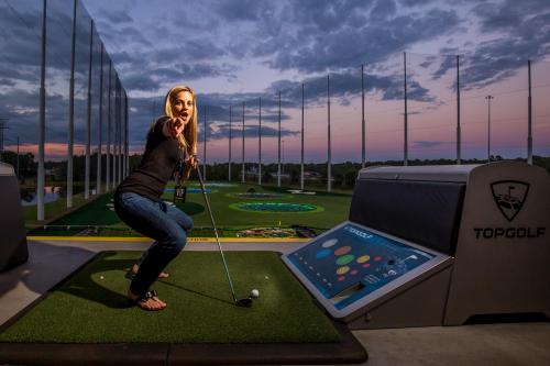 A woman in casual attire prepares to swing a golf club at Topgolf, with a scenic sunset in the background.