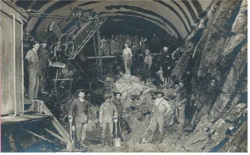 Black and white photo of miners in a tunnel, posing with tools and machinery amidst rock and debris.