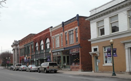 Historic downtown buildings line a street, featuring brick facades and storefronts, with parked cars along the curb.