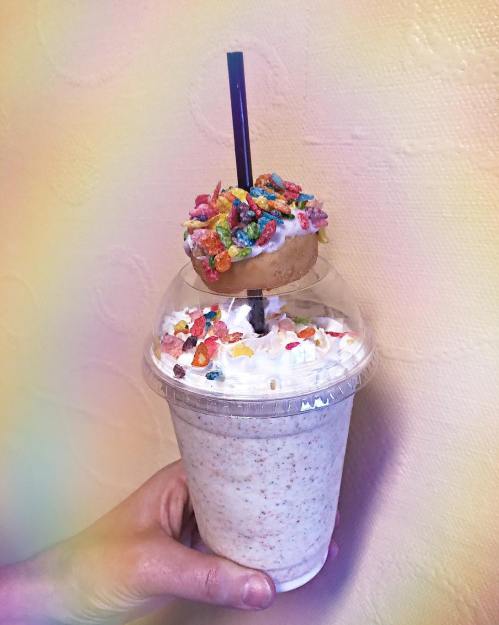 A hand holds a colorful milkshake topped with whipped cream, a donut, and fruity cereal, against a soft pastel background.