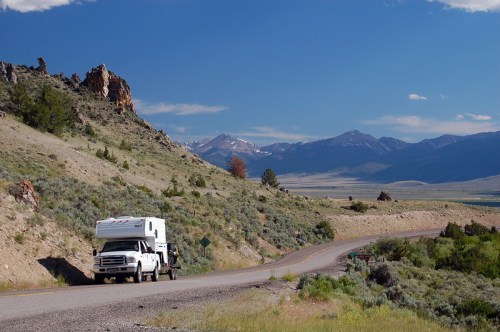 A truck with a camper drives along a winding road surrounded by mountains and greenery under a clear blue sky.