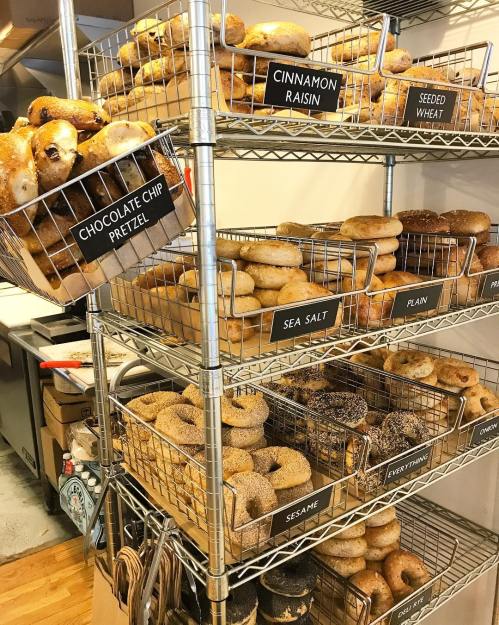 A display of various bagels on wire shelves, labeled with flavors like chocolate chip pretzel, cinnamon raisin, and everything.