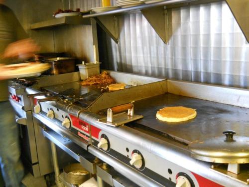 A busy kitchen with a griddle cooking pancakes and other food, with a person serving on the side.