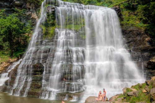 A scenic waterfall cascades over rocky cliffs, with people relaxing on the rocks in the foreground.