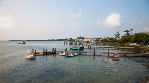 A serene waterfront scene with boats docked at a pier, surrounded by calm water and distant buildings under a cloudy sky.