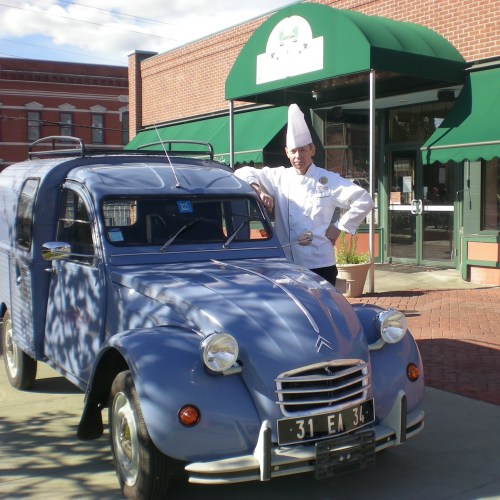 A chef in a white uniform stands next to a vintage blue Citroën car outside a restaurant with green awnings.