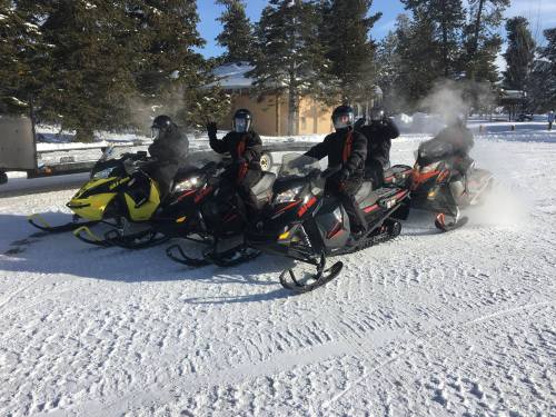 Four snowmobiles parked on snowy ground, with riders in winter gear giving thumbs up against a backdrop of trees.