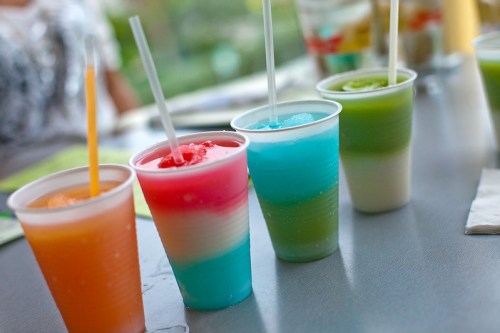 Four colorful layered drinks in plastic cups, featuring orange, pink, blue, and green hues, with straws.
