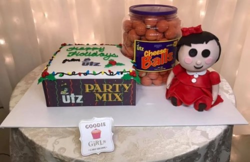 A festive table with a holiday-themed cake, Utz party mix, cheese balls, and a doll in a red dress.