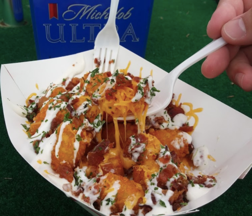 A hand holds a fork over a tray of loaded tater tots topped with cheese, bacon, and drizzled with ranch dressing.
