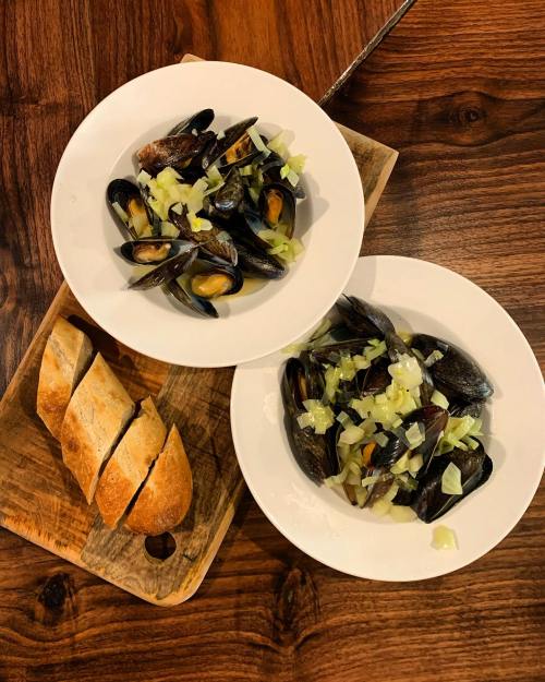 Two bowls of mussels with chopped onions, served with a sliced baguette on a wooden board.