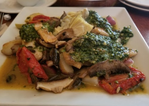 A plate of grilled fish topped with green sauce, surrounded by roasted vegetables and artichokes.
