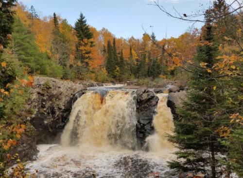 A waterfall cascades over rocks, surrounded by vibrant autumn foliage and evergreen trees.
