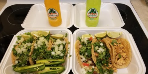 Two takeout containers with tacos topped with onions, cilantro, and jalapeños, alongside bottles of Jarritos soda.