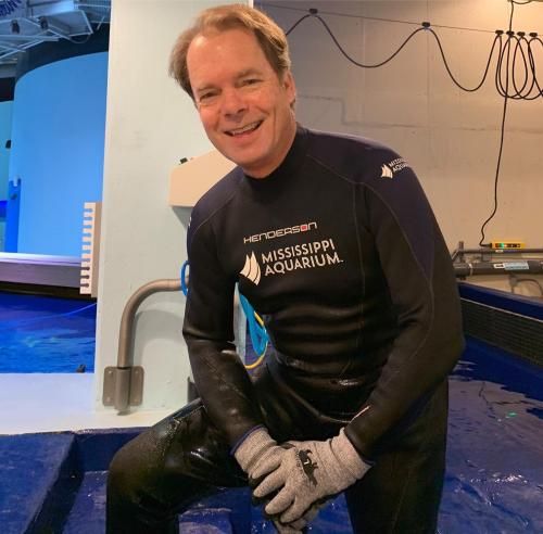 A man in a wetsuit and gloves smiles while sitting on the edge of a pool at the Mississippi Aquarium.