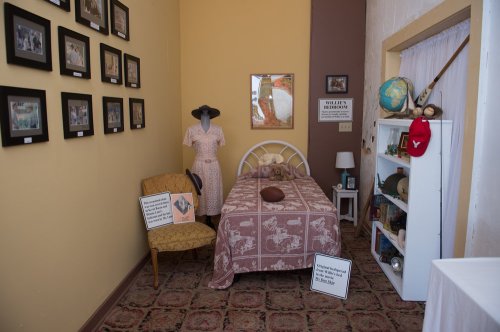 A small room featuring a bed, chair, vintage dress, and framed pictures on the wall, with informational signs displayed.