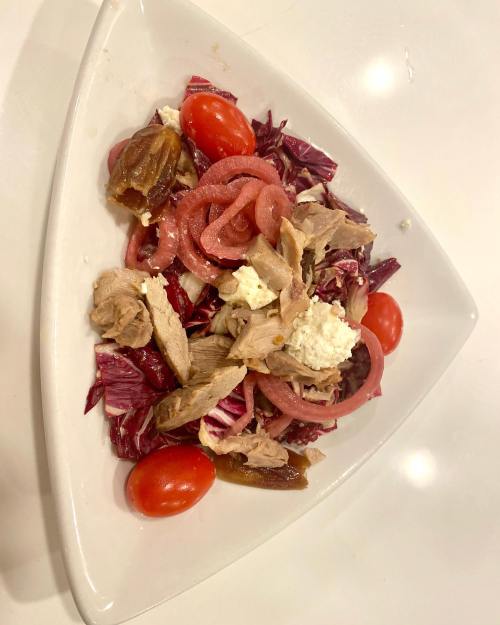 A triangular plate with a salad featuring shredded chicken, cherry tomatoes, pickled onions, dates, and cheese on greens.