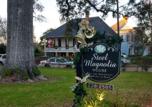 Sign for Steel Magnolia House Bed and Breakfast, surrounded by greenery and festive decorations, with a historic home in the background.