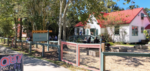 A charming restaurant with a red-roofed building, outdoor seating, and a welcoming sign, surrounded by trees.