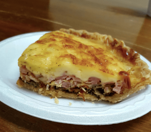 A slice of savory pie with layers of ham, cheese, and a golden crust on a white paper plate.