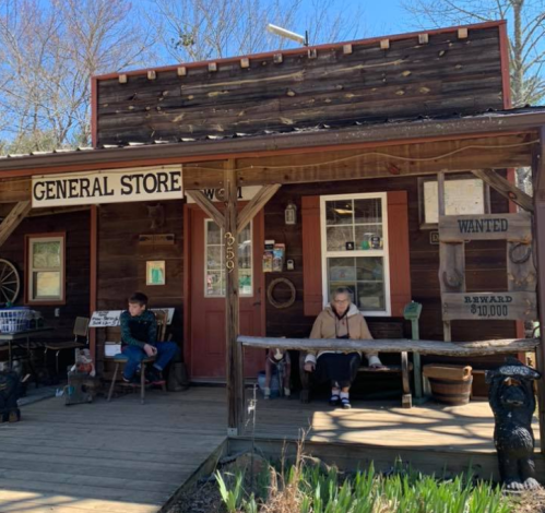 A rustic general store with wooden exterior, featuring two people sitting on the porch and a "Wanted" sign nearby.