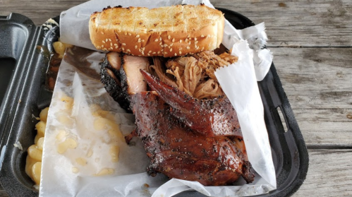 A takeout container with barbecue meat, a slice of bread, and macaroni on a wooden table.