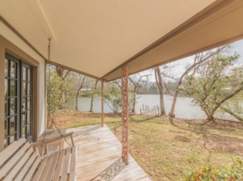 A porch swing overlooks a serene lake surrounded by trees on a sunny day.