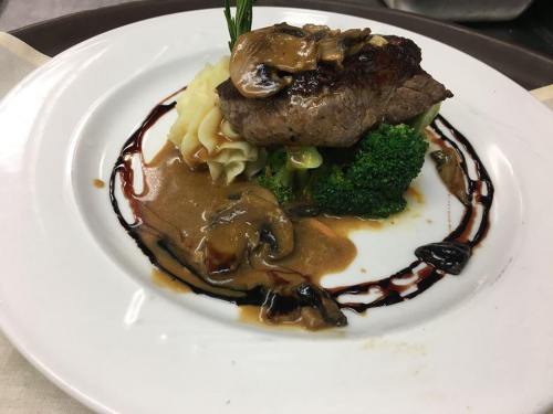A plated dish featuring steak topped with mushrooms, served with mashed potatoes and broccoli, drizzled with sauce.
