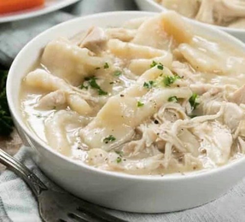A bowl of chicken and dumplings in creamy broth, garnished with parsley, served with a side of carrots.