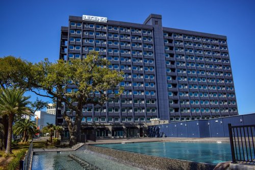 A modern, dark blue hotel building with large windows, surrounded by palm trees and a water feature in the foreground.