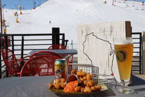 A plate of cheese curds and a beer sit on a table with a snowy ski slope in the background.