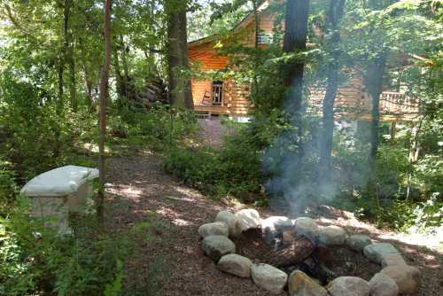 A cozy cabin surrounded by trees, with a stone fire pit and smoke rising from a small campfire.