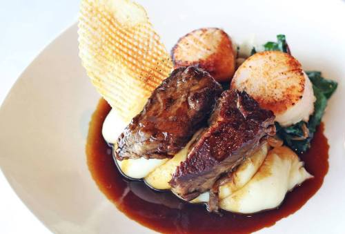 A gourmet dish featuring tender meat, scallops, creamy mashed potatoes, and a crispy garnish, drizzled with sauce.