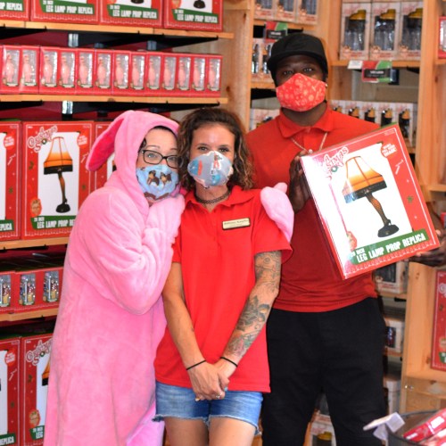 Three people pose in a store, one in a pink onesie, another in a red shirt, and a third in a mask, surrounded by festive decorations.