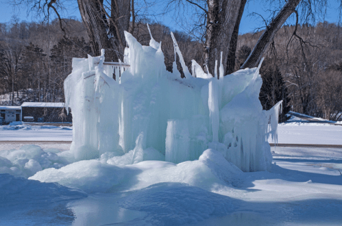 A frozen ice formation with sharp icicles, surrounded by snow and trees under a clear blue sky.