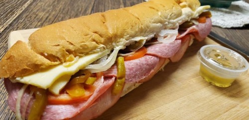 A large sub sandwich filled with meats, cheese, and vegetables, served with a small container of dressing on a wooden board.
