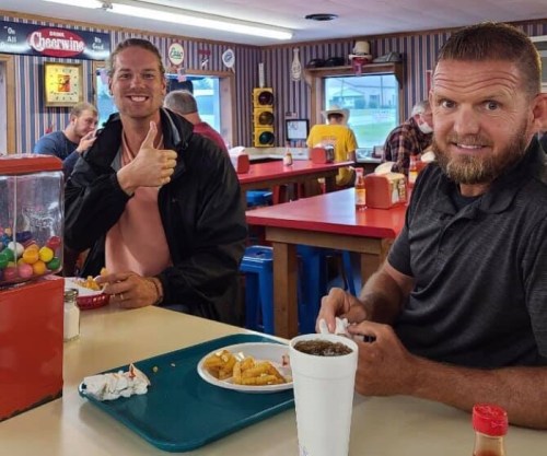 Two men sitting at a diner table, smiling and enjoying food, with a gumball machine in the background.