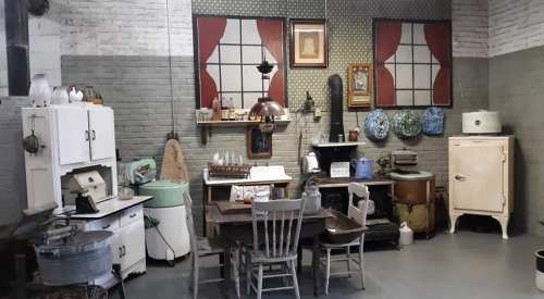 A vintage kitchen scene featuring old appliances, a wooden table, chairs, and decorative wall art.